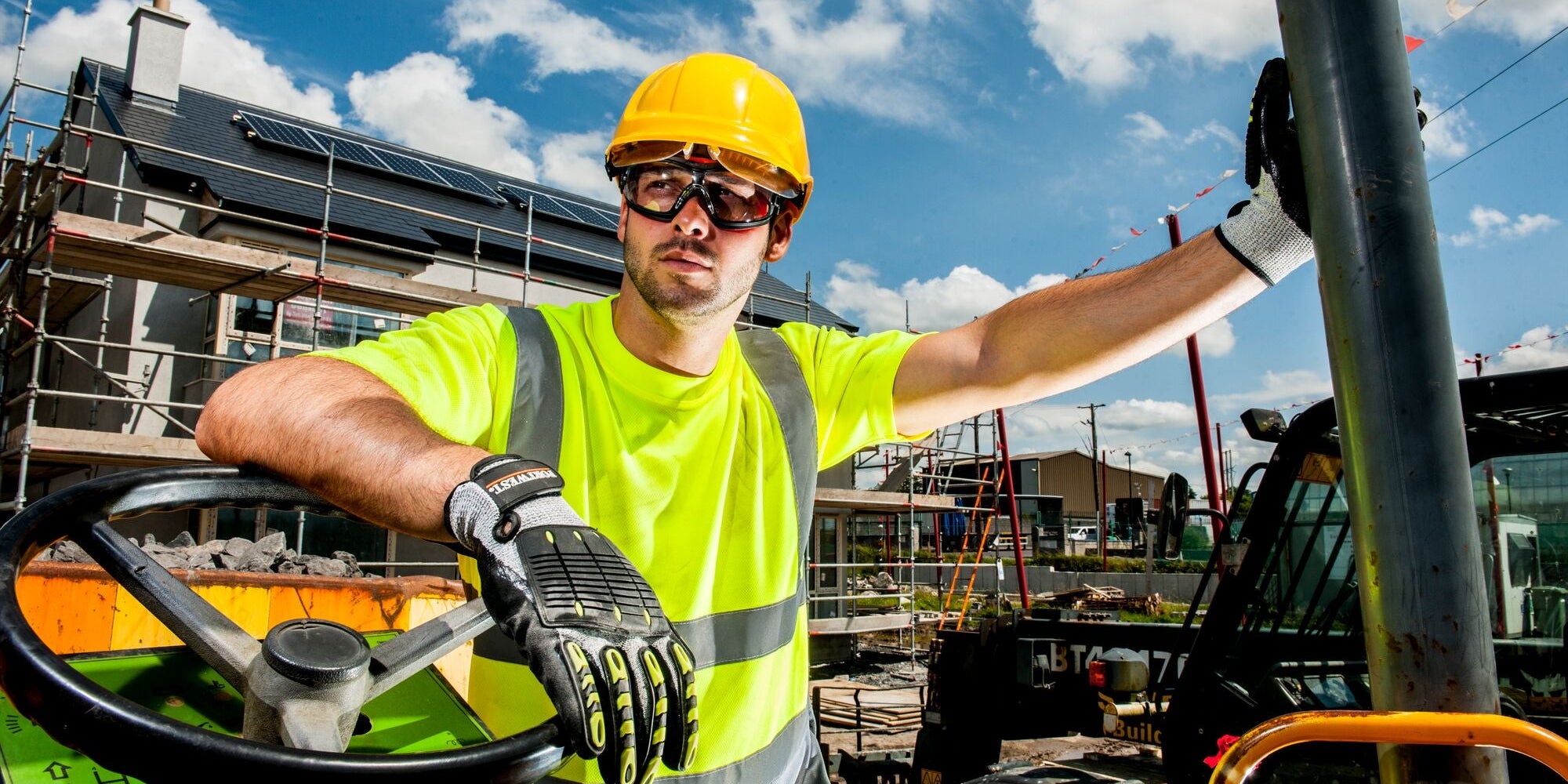 Worker in hi-vis T-shirt, safety helmet, goggles, and gloves leaning on forklift in a work construction site
