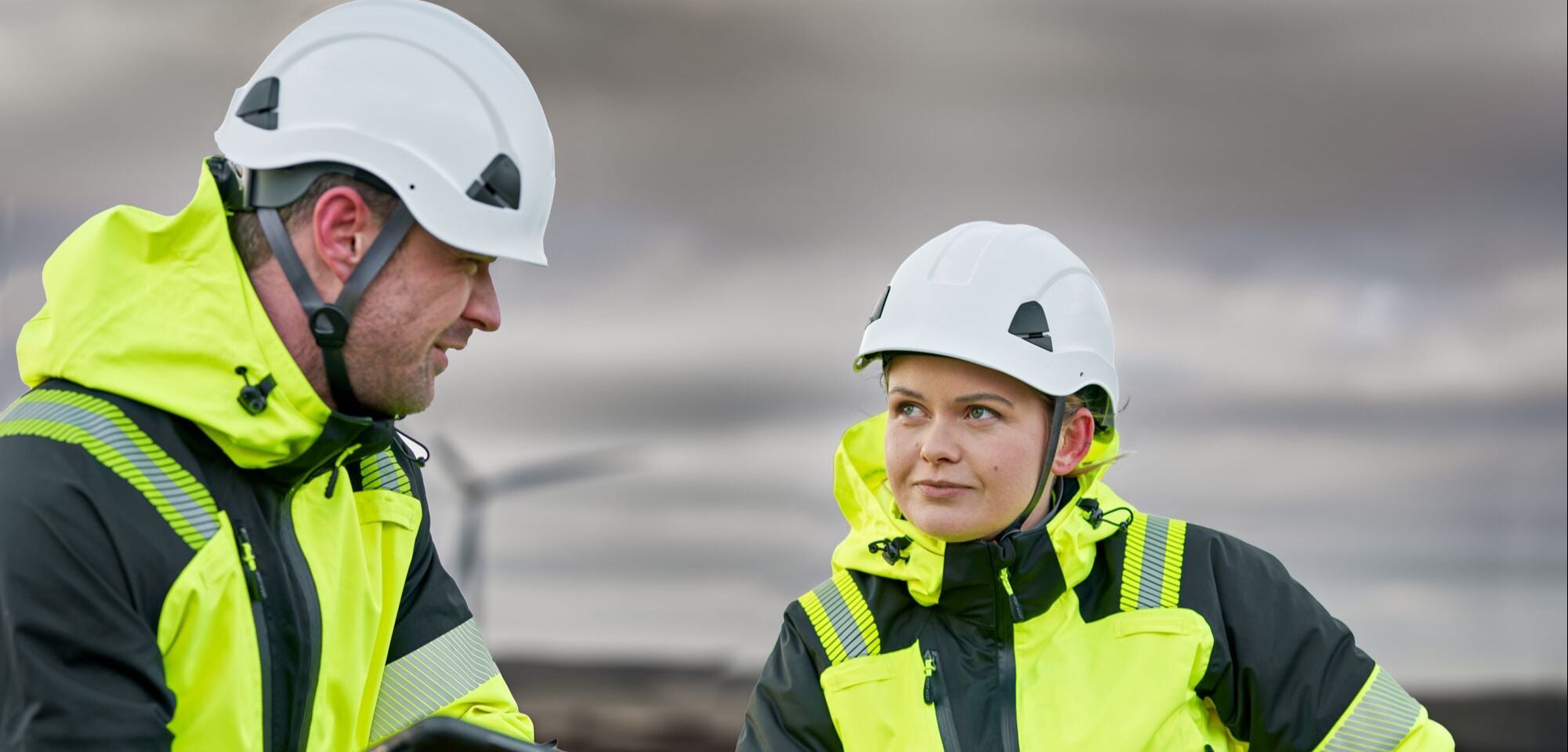 Two individuals in high-visibility jackets and white helmets, sitting outdoors with a blurred background.