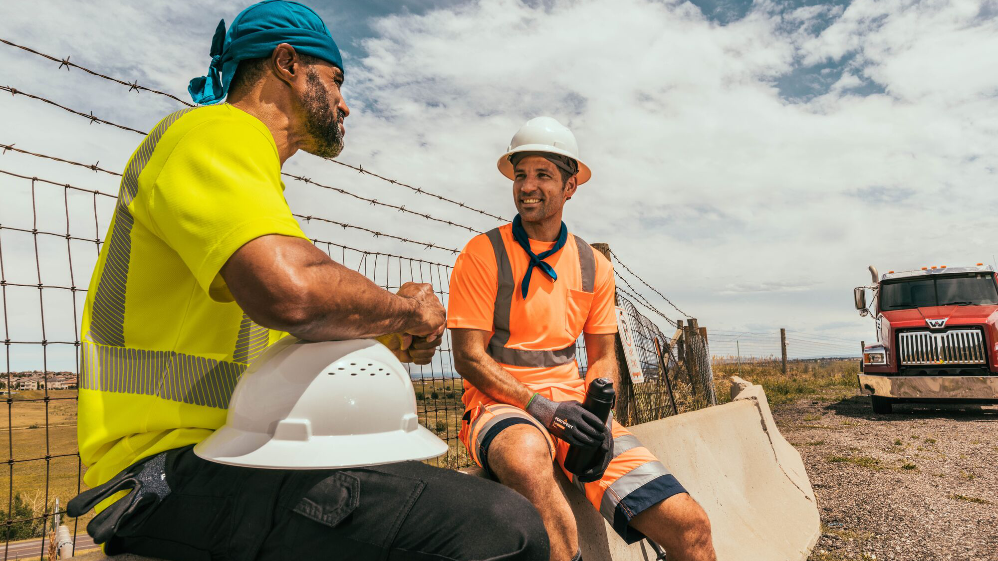 Two men wearing hi-vis short sleeve t-shirts one in neon yellow with segmented reflective tape, and the other in bright orange with silver reflective tape both seated and taking a break at a construction site under a partly cloudy sky by MAK Boots & Garments.