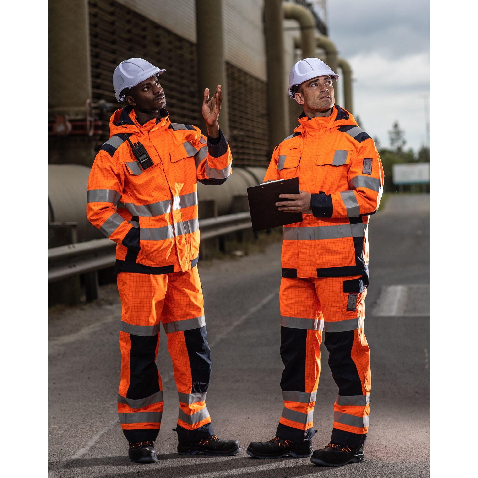Two workers wearing orange flame-resistant high-visibility jackets with reflective tape and white hard hats, communicating on a worksite with one using a radio and the other holding a clipboard. Designed for enhanced safety in hazardous environments. By MAK Boots & Garments.