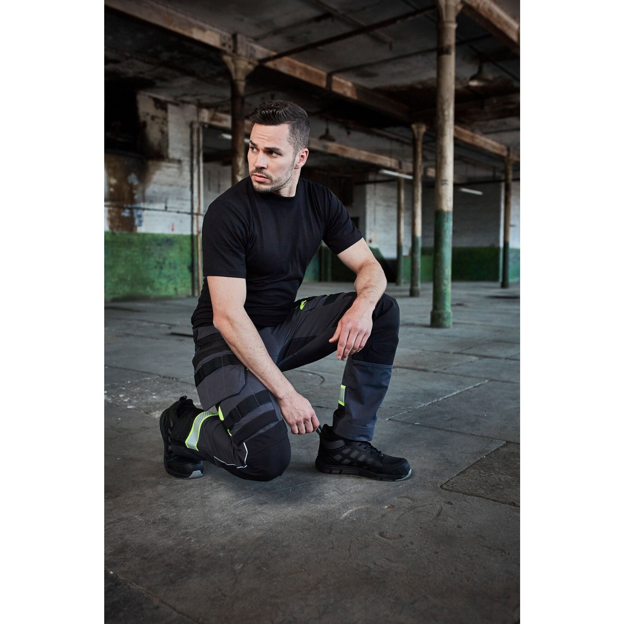 Worker kneeling in rugged black work trousers with reinforced knee panels and multiple pockets, paired with durable tan safety boots. Built for strength, flexibility, and utility on the job. By MAK Boots & Garments.